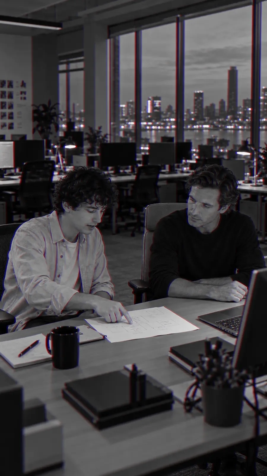 Two men sit together at a desk in an empty office after dark, city lights behind them, working through a document.