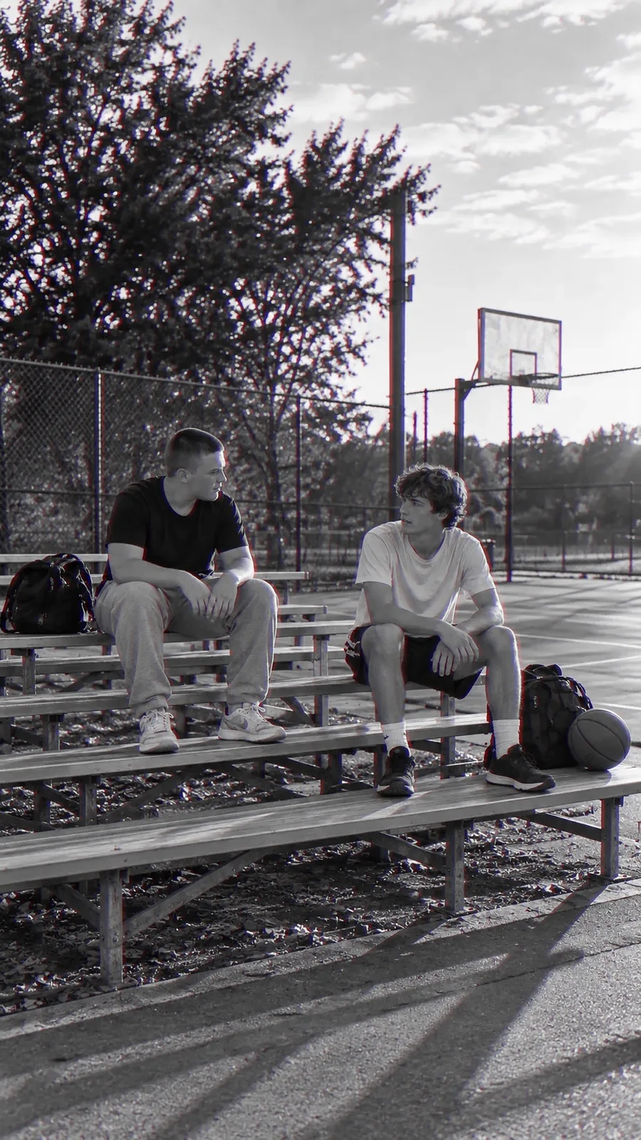 Two young men sit on the bleachers beside a basketball court, one talking and the other listening.