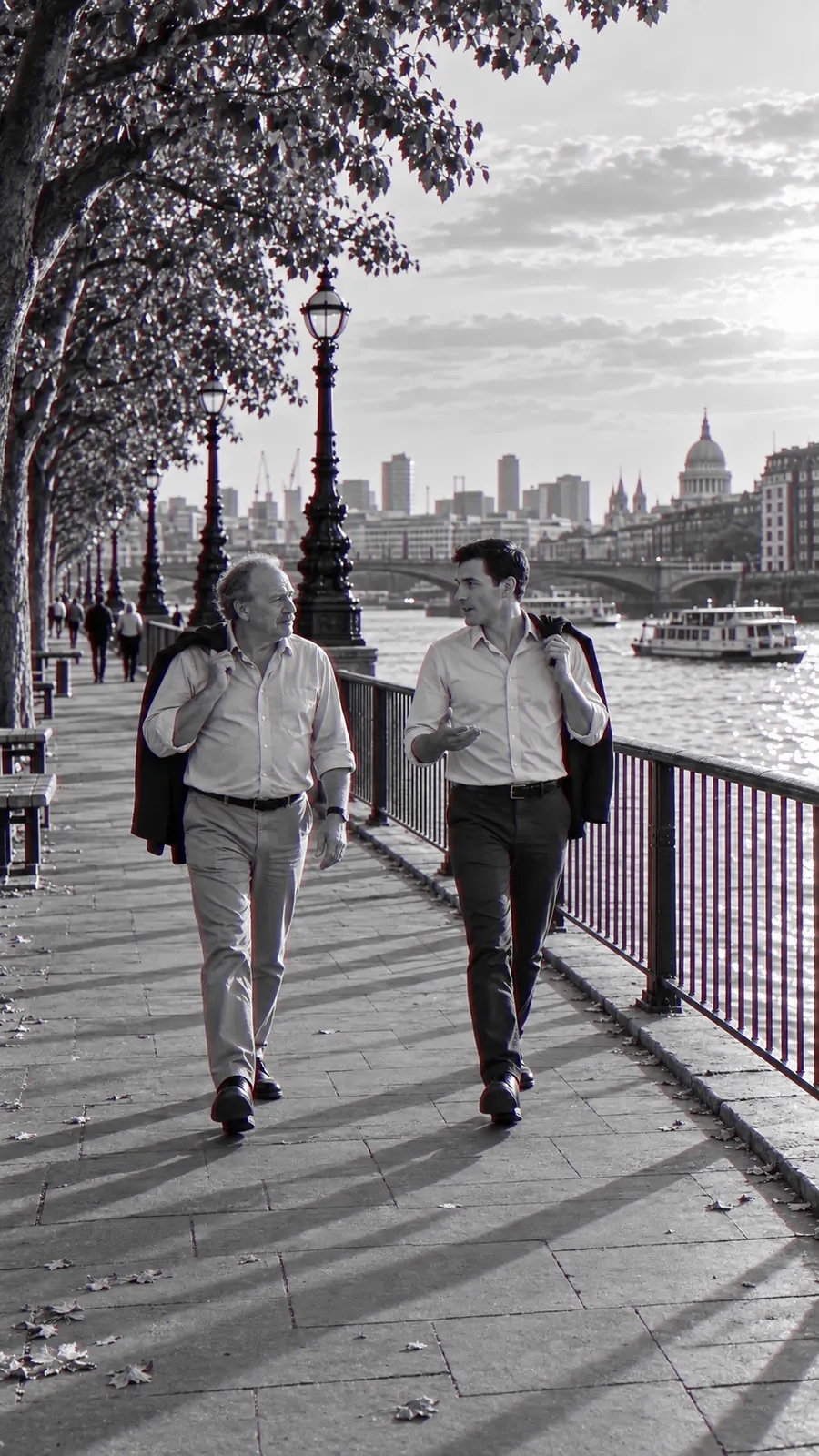 Two men walk along a river path with their jackets over their shoulders, talking something through side by side.
