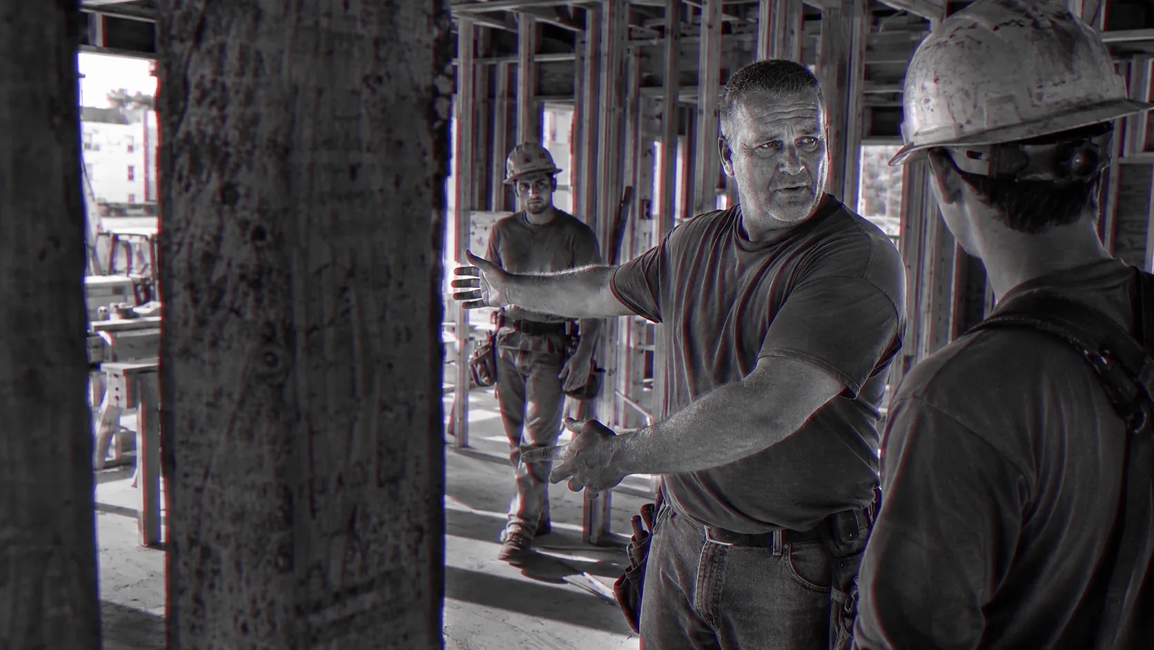 A foreman on a framing site raises a hand to stop the work while two younger men listen.