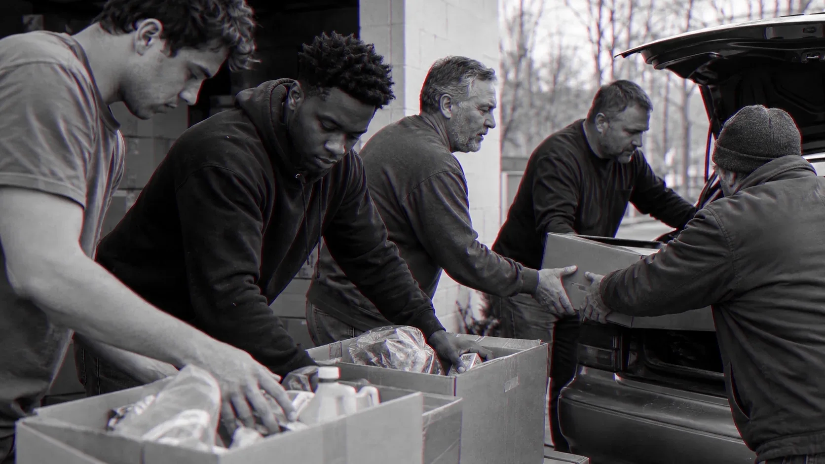 Four men load boxes of food into a car boot for delivery, the youngest lifting first.