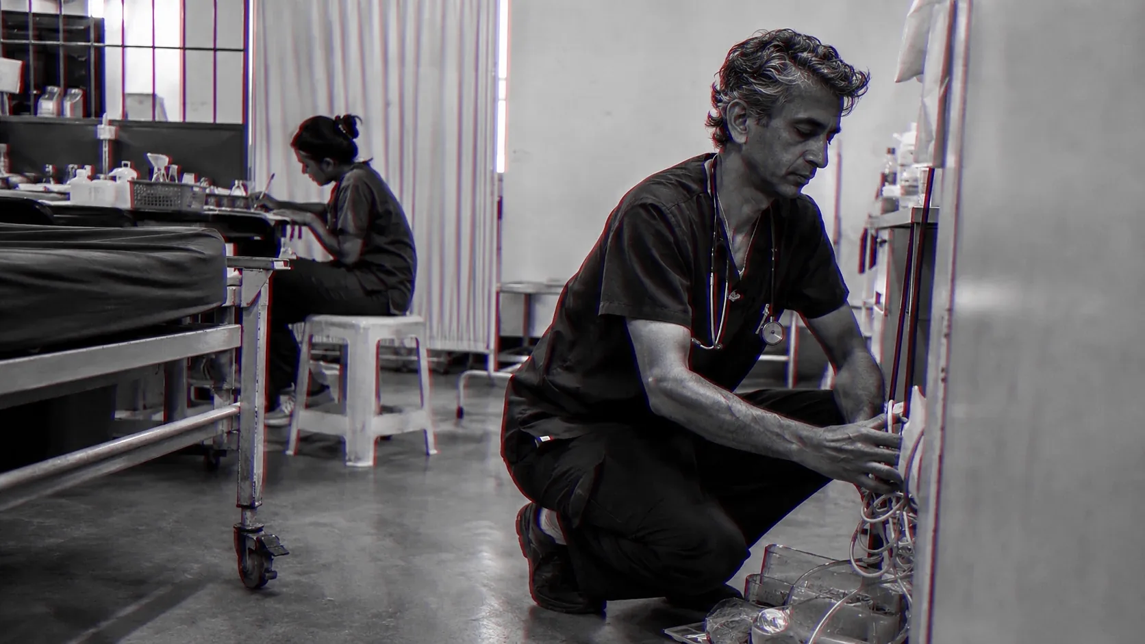 A physician kneels to clean and reset a clinic after the last patient has gone, while a colleague works behind him.