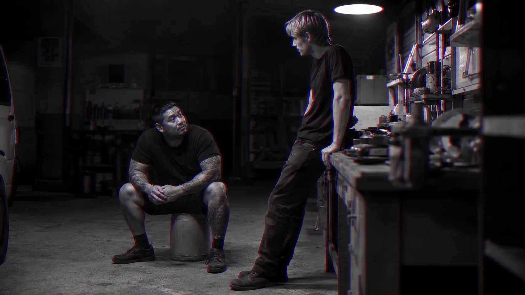 One man sits on an upturned crate in a workshop while another leans on the bench and listens.