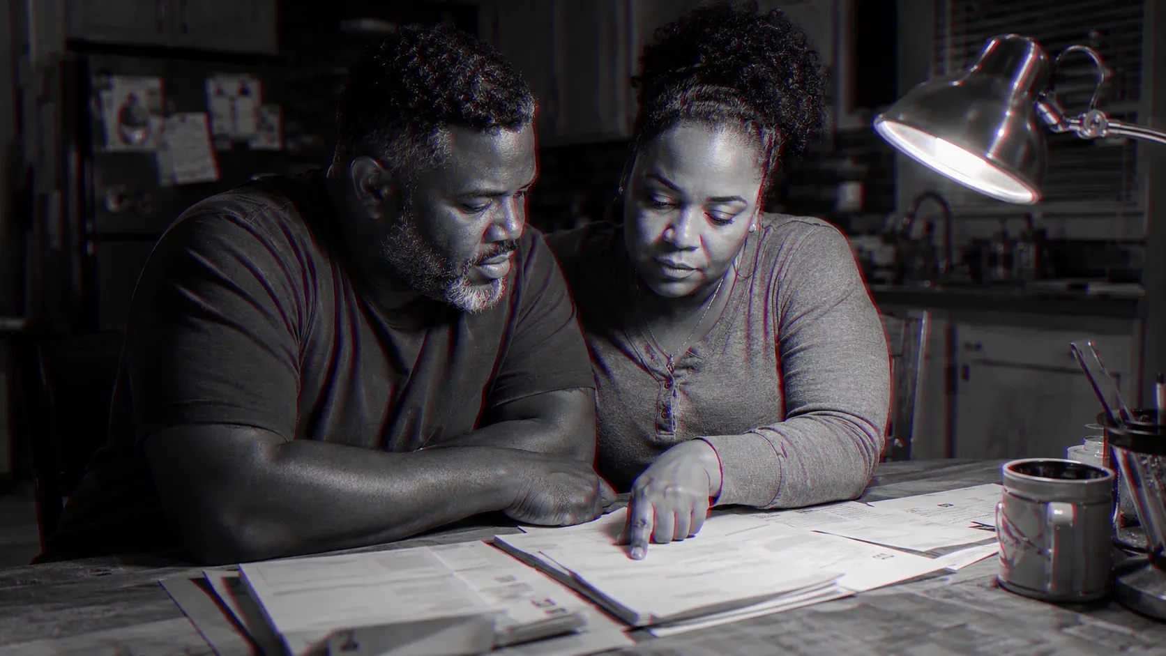 A man and a woman sit side by side at a table at night, working through household papers as equals.