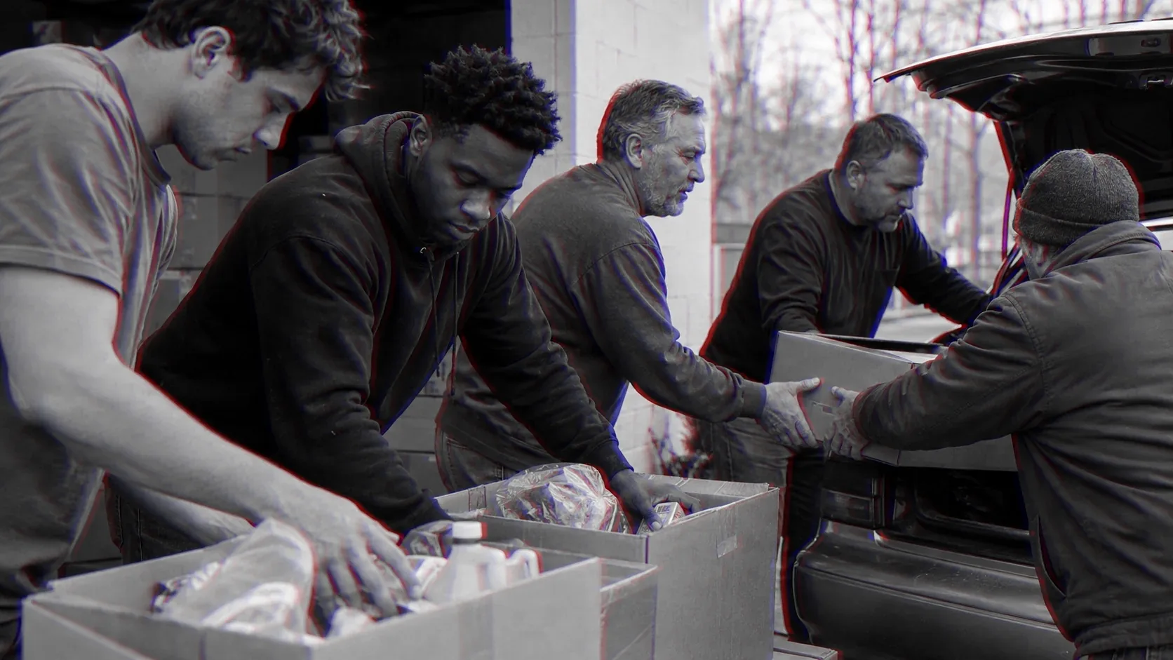 Four men load boxes of food into a car boot for delivery, the youngest lifting first.