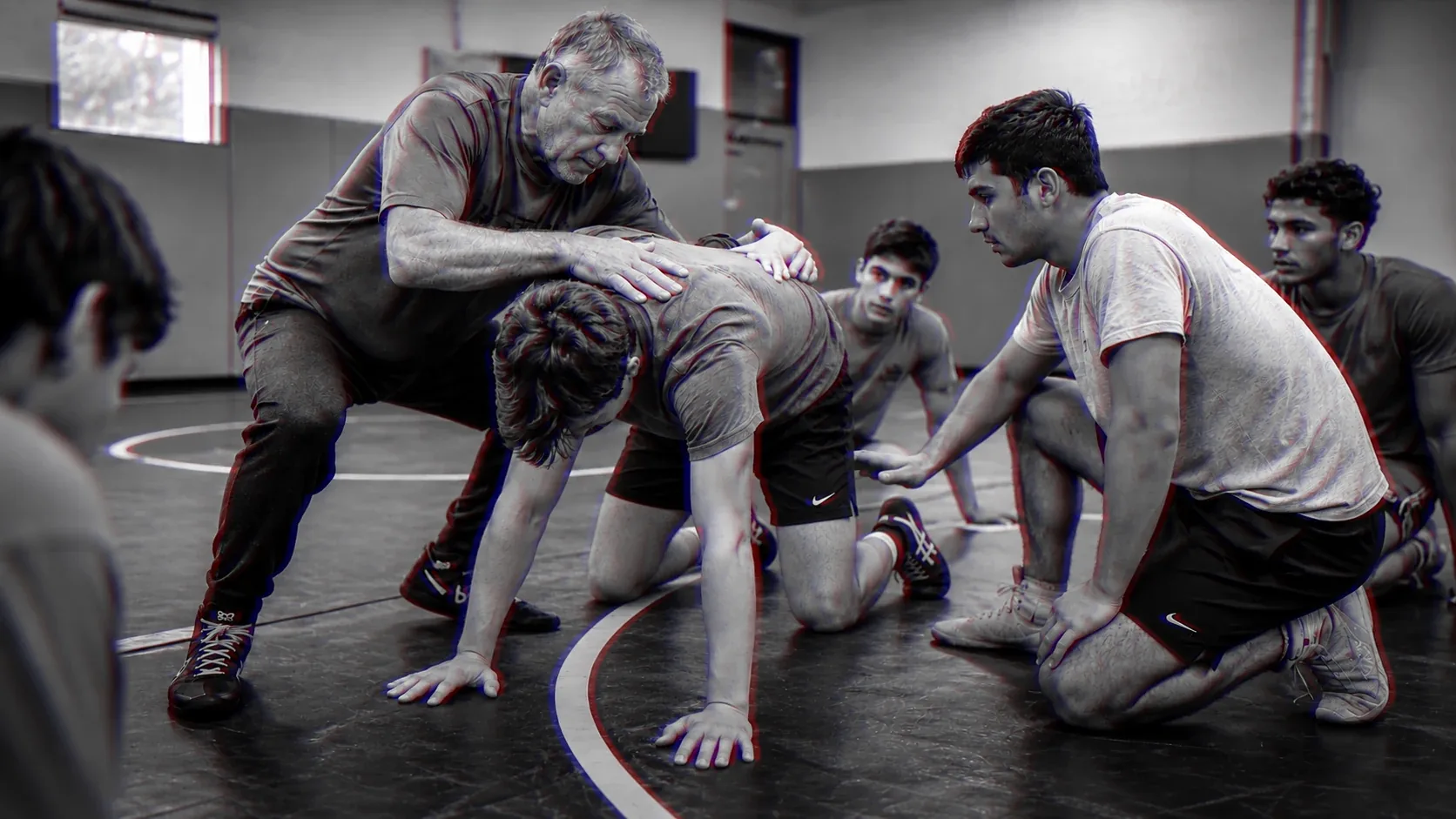 A coach sets a young wrestler’s stance while four teammates kneel around the mat and watch the correction.