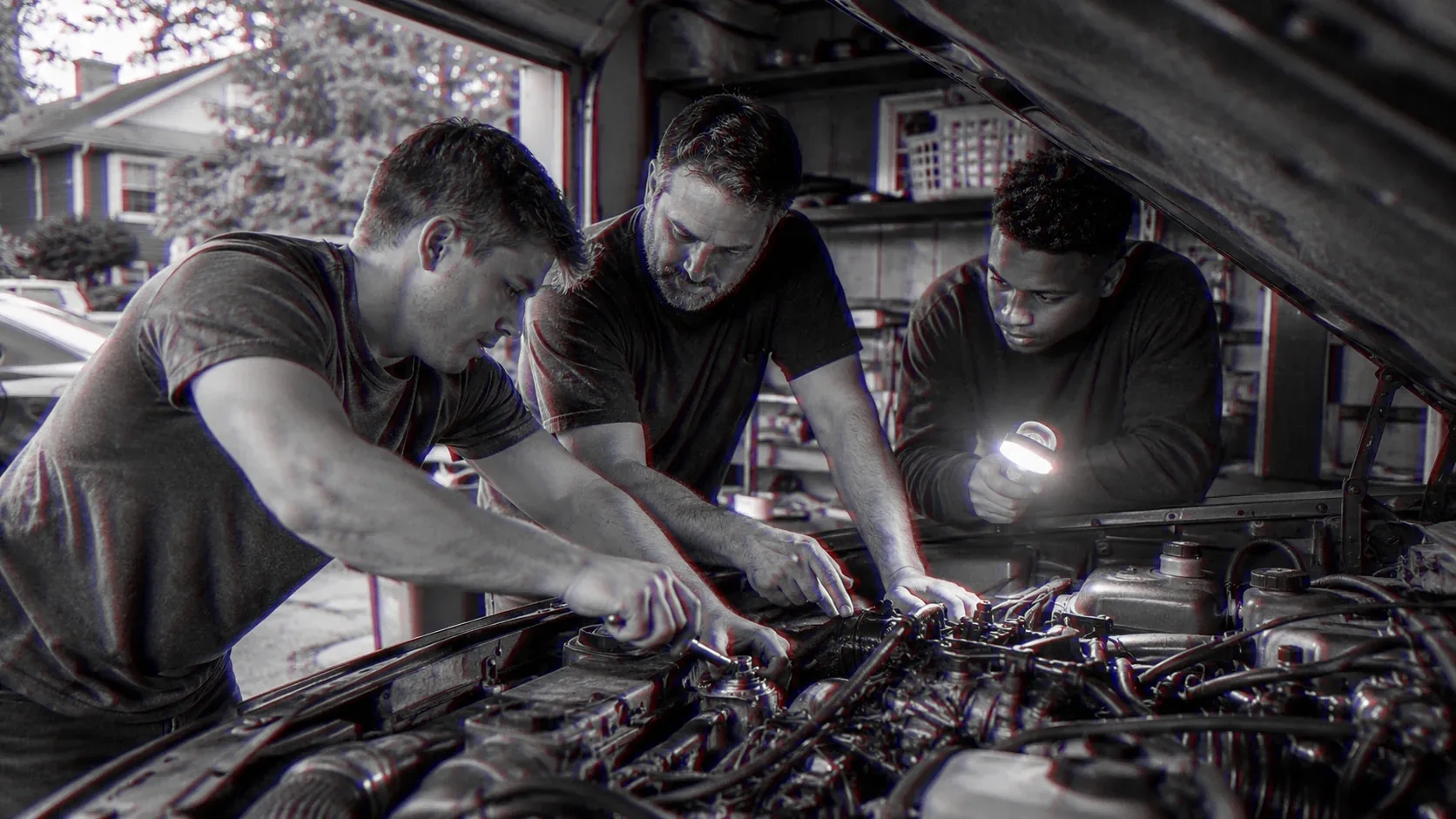 Three men work an engine bay by lamplight, an older man guiding a younger one’s hands.