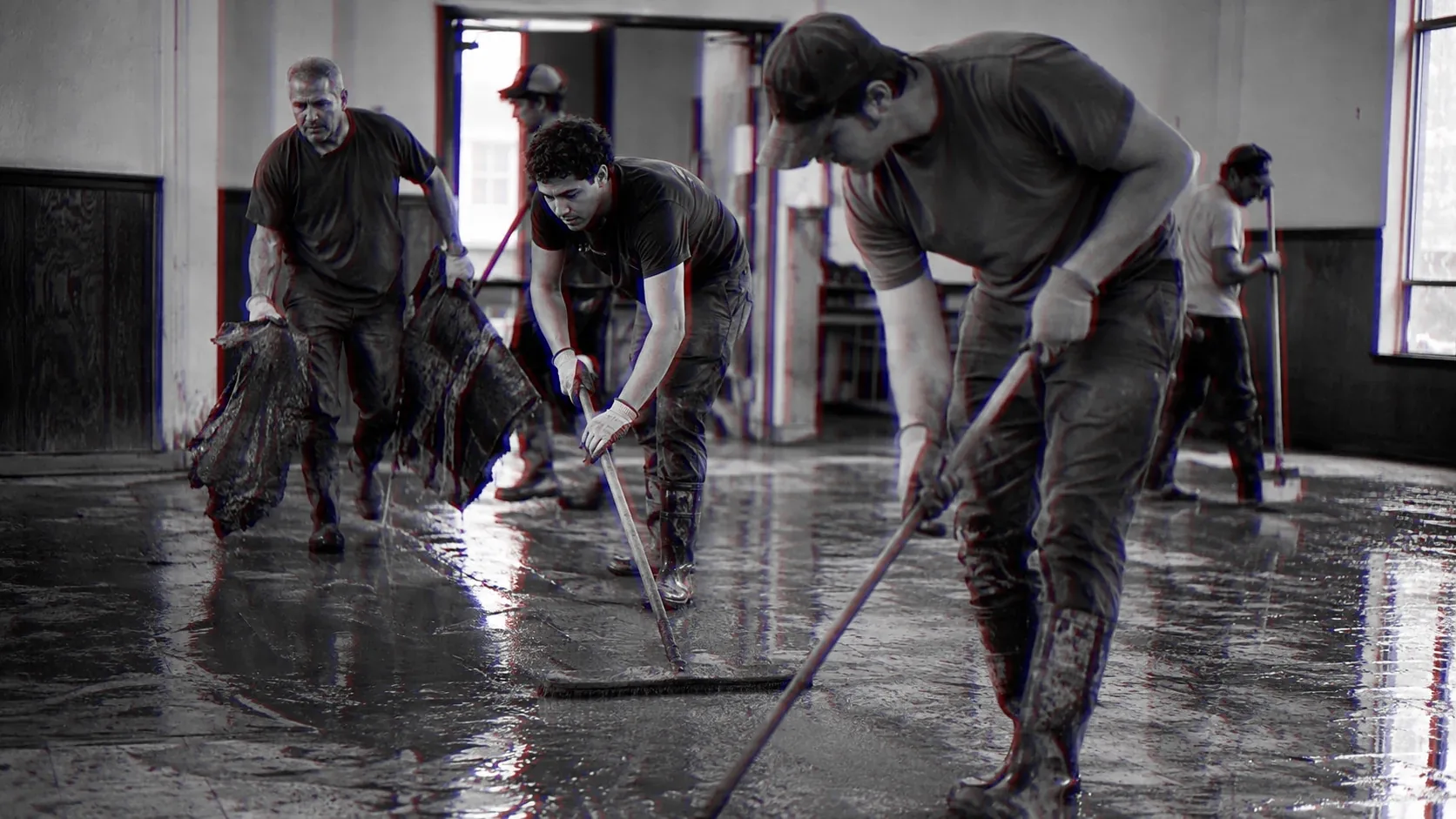 Three men push water out of a flooded hall with squeegees, boots soaked.