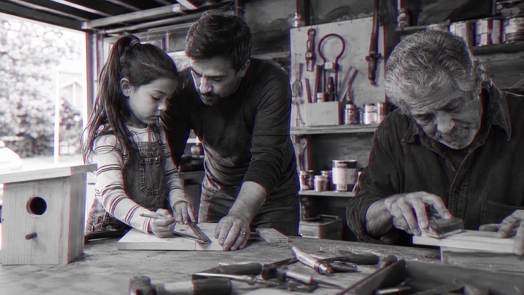 A father guides his daughter’s hands on a birdhouse at a workbench, an older man working behind them.
